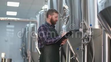 有胡子的男酿酒师布鲁塔站在啤酒罐附近，记录仪器读数。 手工艺啤酒生产